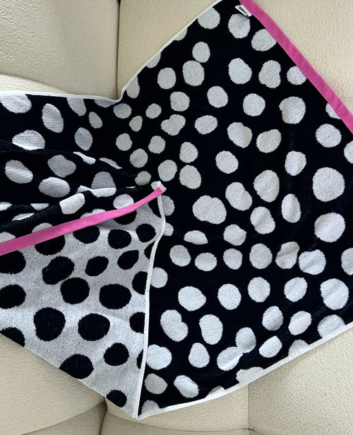 A square black and white polka dot towel with a bright pink trim, laid out on a cream-colored leather chair.