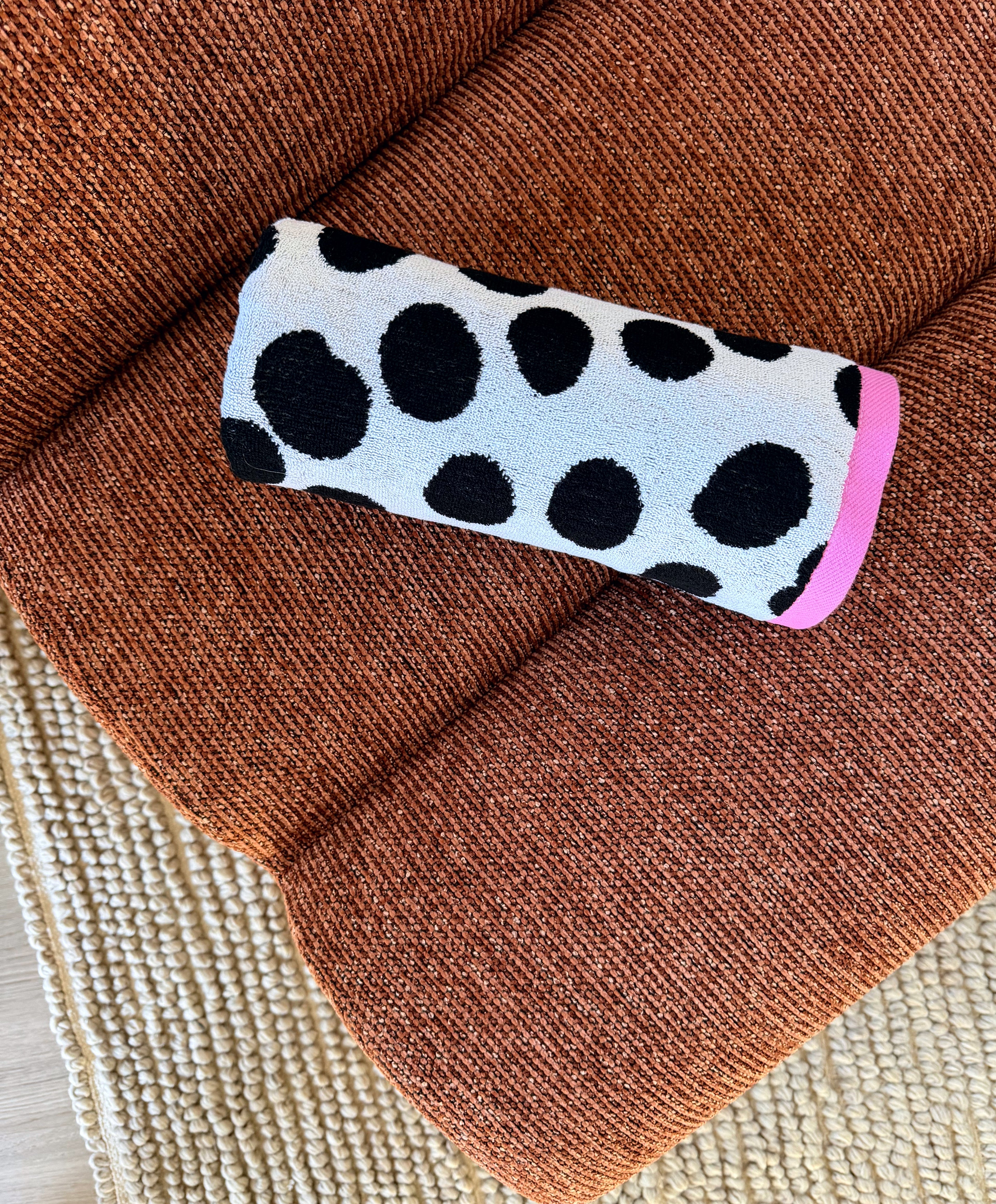 A cylindrical fabric item, likely a pet lick mat or small cushion, featuring a black and white cow print pattern with a bright pink end cap, resting on a textured rust-brown upholstered chair.